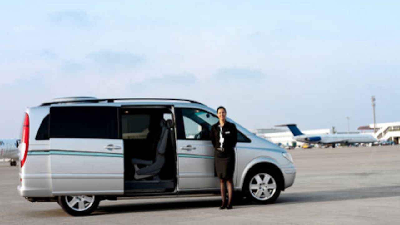 Alt text: A woman stands beside a van on the airport tarmac, with planes and terminal buildings visible in the background. 
