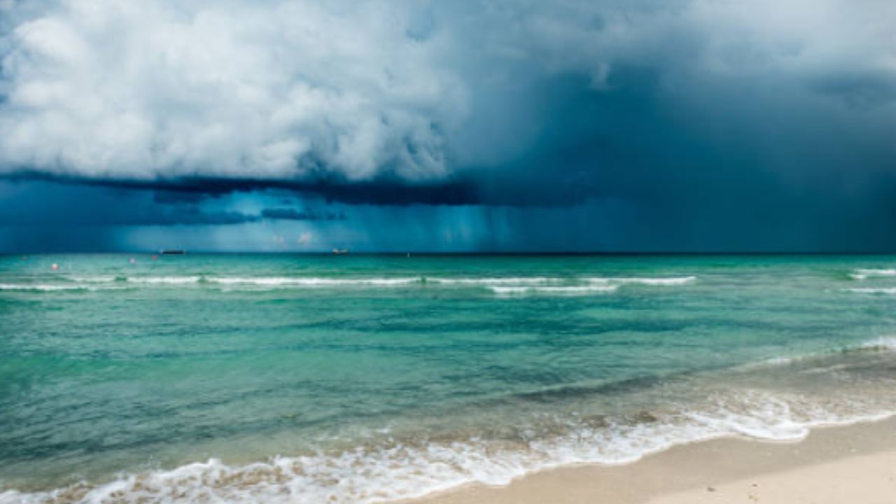 Dark storm clouds gather over a turbulent ocean, casting shadows on a deserted beach below.