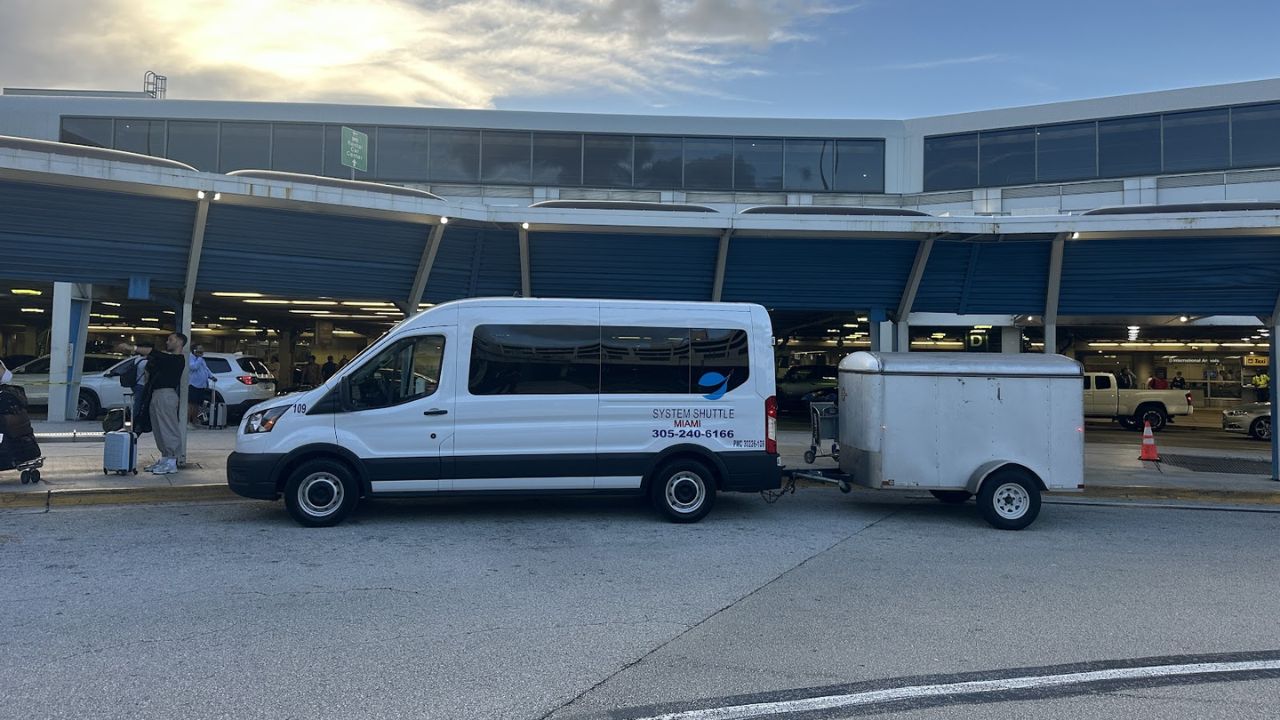 System Shuttle Miami van parked at airport terminal with luggage trailer attached.