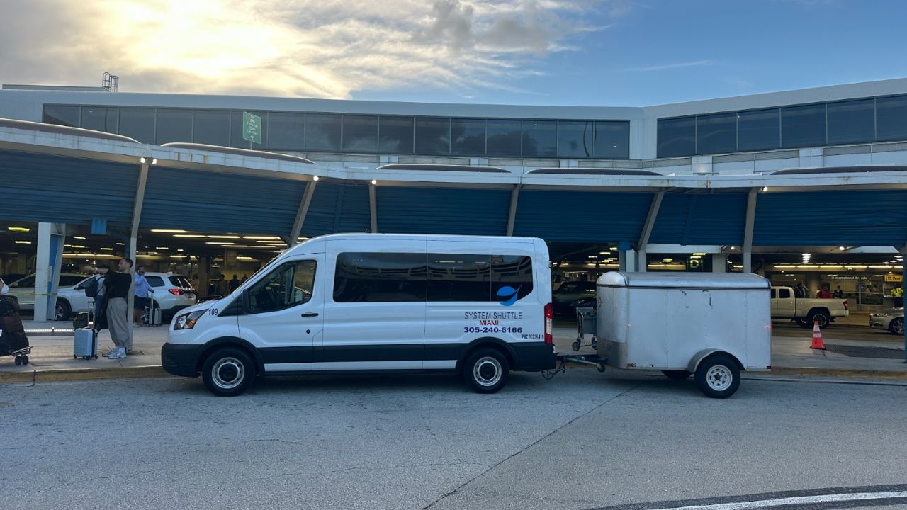 System Shuttle Miami white passenger van towing a small enclosed trailer at an airport curbside pickup area