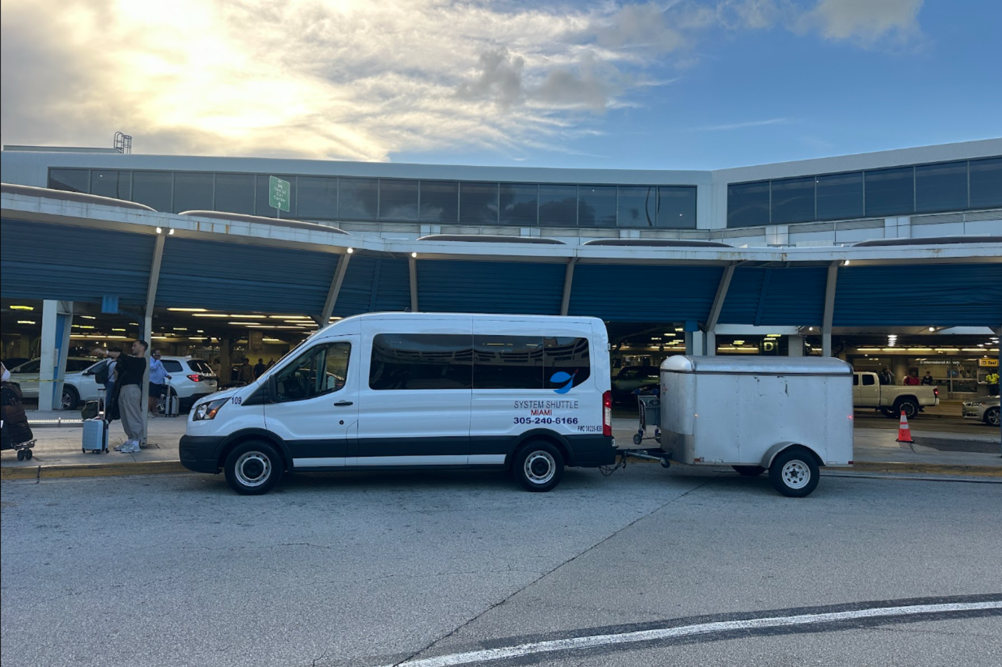 System Shuttle Miami airport shuttle van with luggage trailer waiting outside Miami International Airport for passenger pickup and transfers.