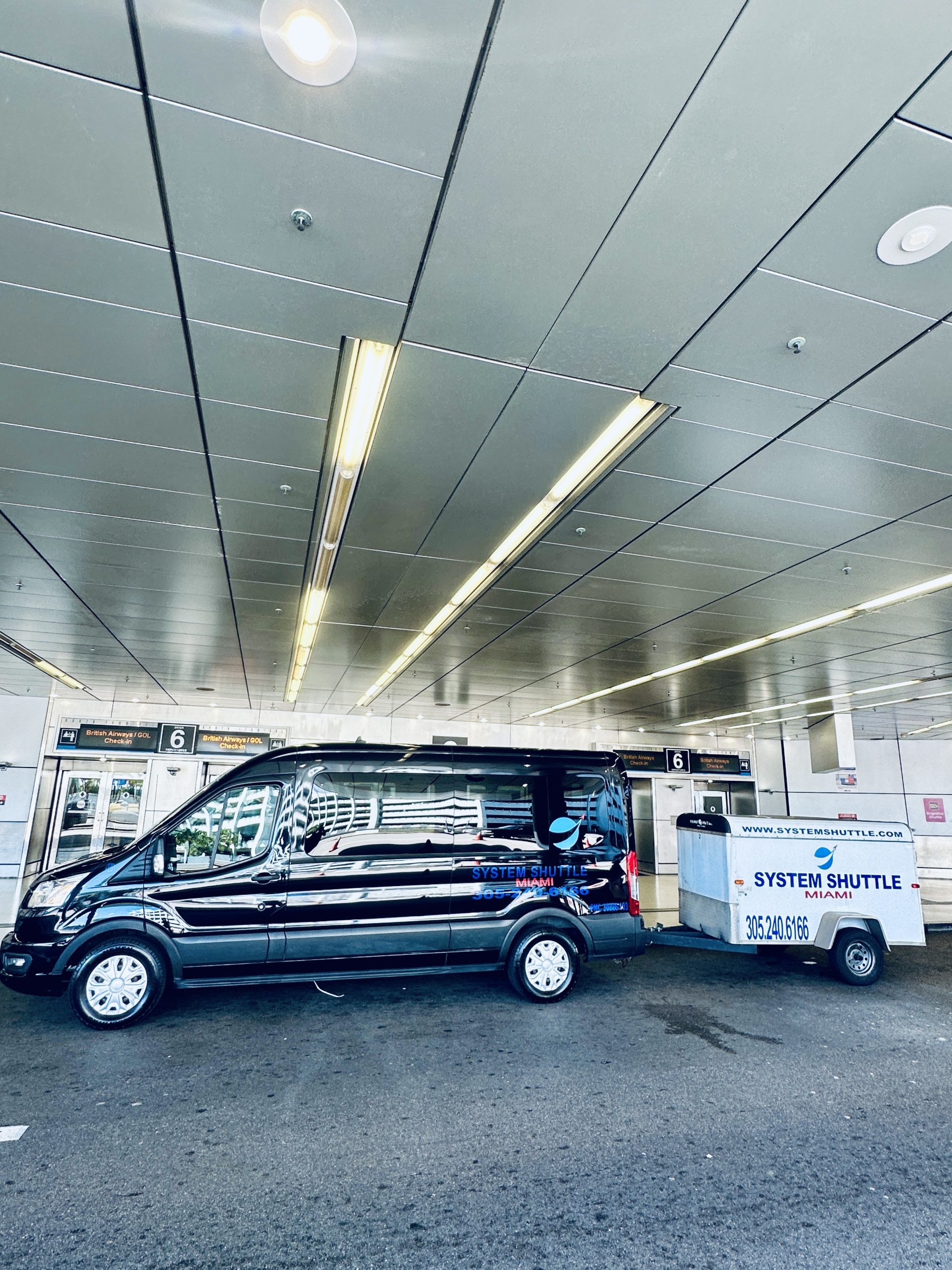System Shuttle Miami airport shuttle van towing luggage trailer at pickup terminal.