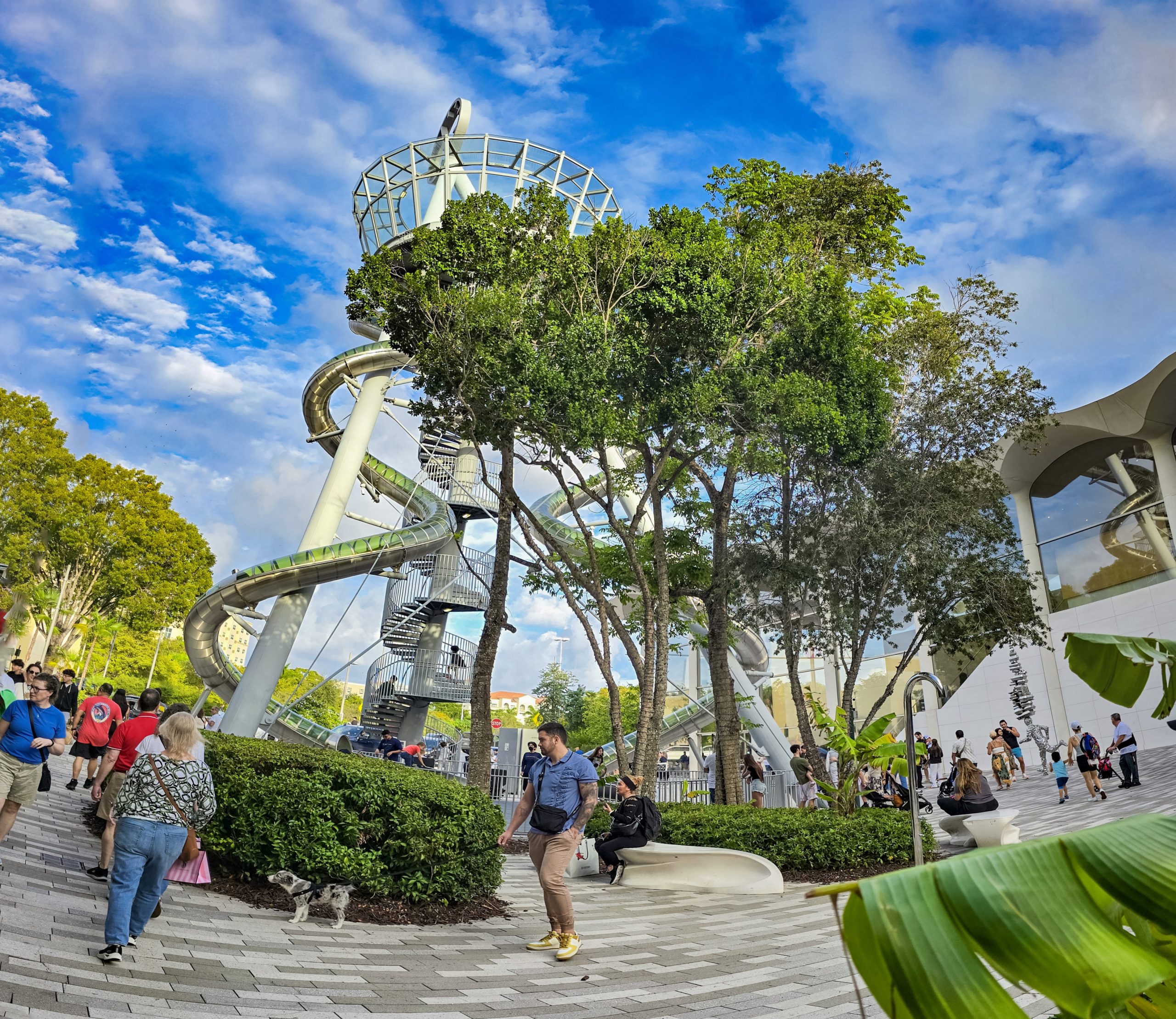 Tall outdoor spiral slide structure surrounded by trees and pedestrians in an urban plaza.